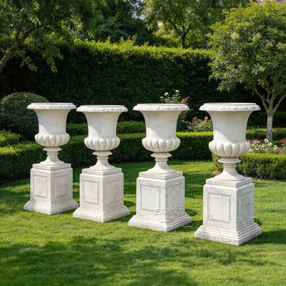 Four white marble flower pots with square pedestals arranged in a garden