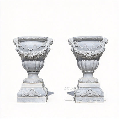 Two white marble relief urn planters with floral garlands and carved bases on a white background